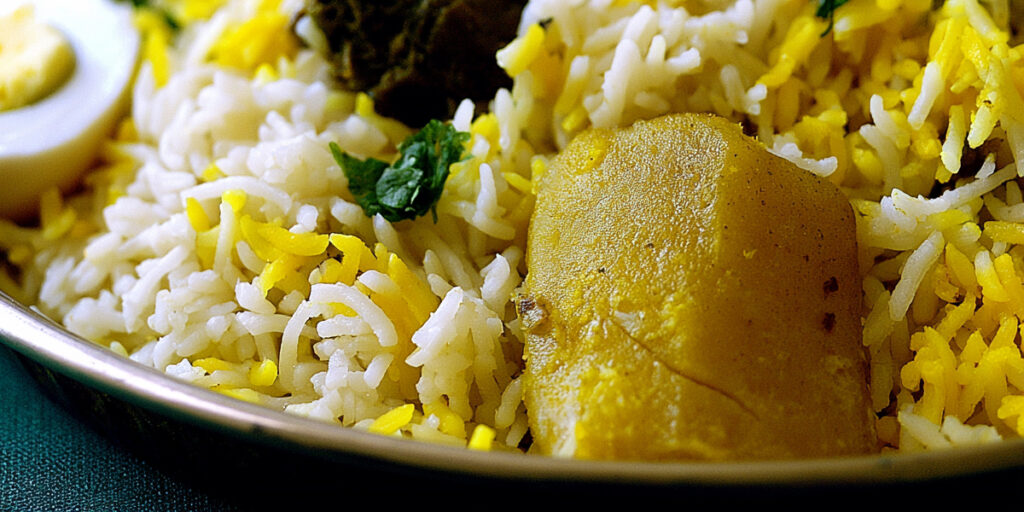 A whole turmeric-stained potato sitting in white basmati rice streaked with saffron, alongside boiled egg and mutton, in a Bengali biryani.